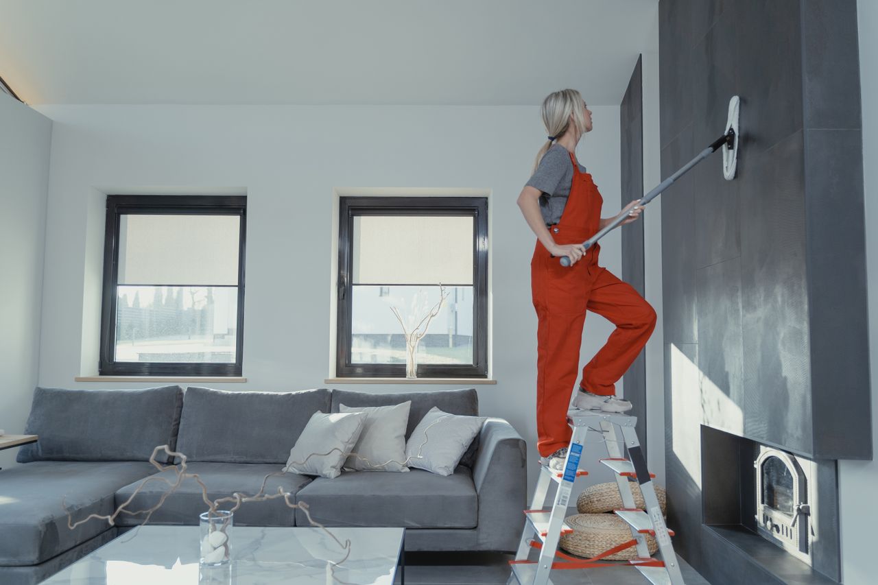 team-02 A woman in overalls is cleaning a wall in a stylish living room, helping maintain a healthy home environment.